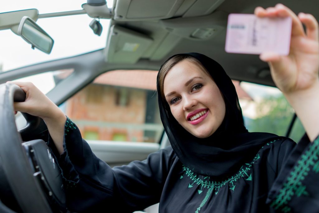 Renewing a driving license in Dubai; happy Muslim woman driving car and holding up Dubai driving license