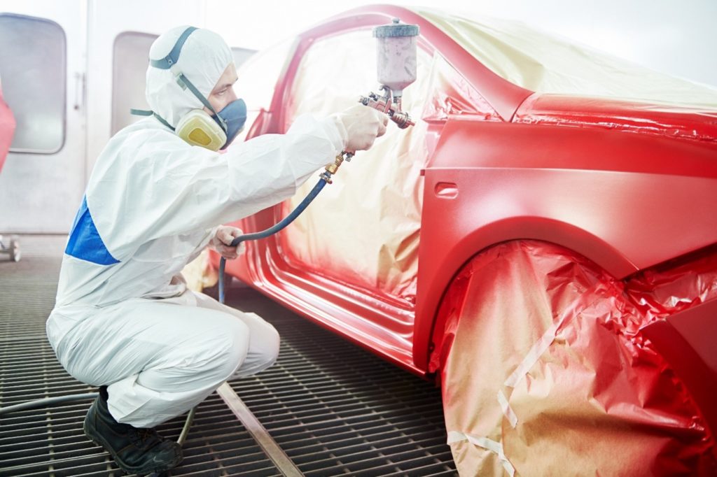 An auto mechanic worker painting a red car in a paint chamber shows how to repaint a car.